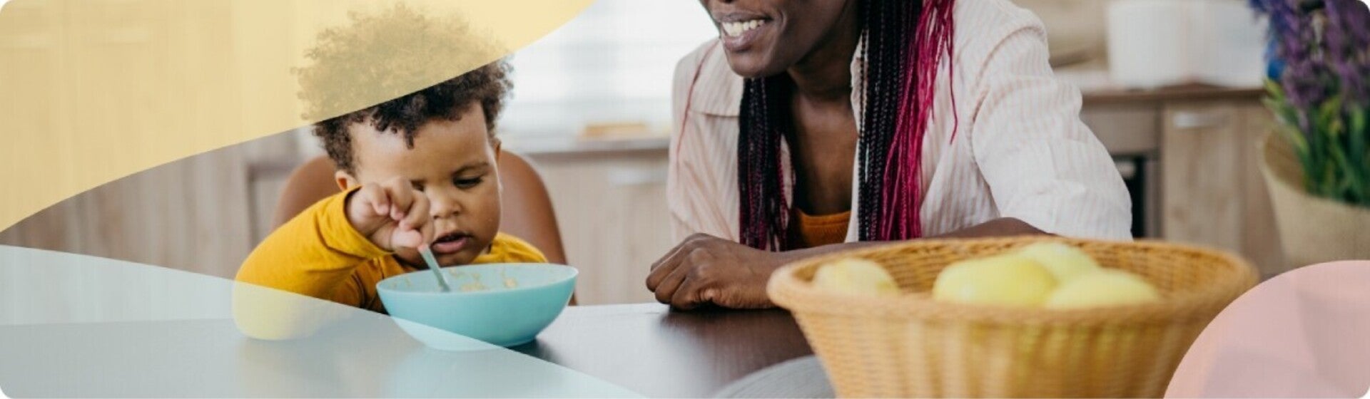 Toddler eating from a bowl with an adult smiling nearby.